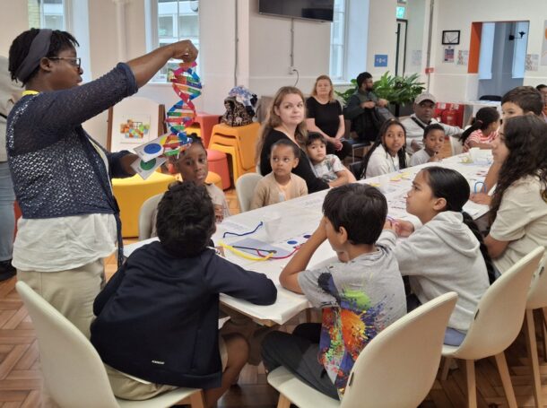 Parents and children seated round a table follow a demonstration by a Regent's Park Time Bank programme leader